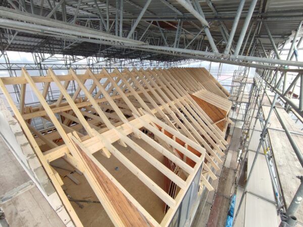 Holz-Dachstuhl eines Gebäudes im Bau, umgeben von einem großen Gerüst mit Schutzdach.